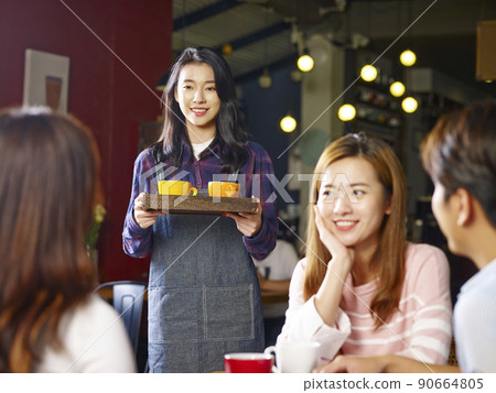 young smiling asian waitress serving customers in coffee shop, focus on the background girl 90664805