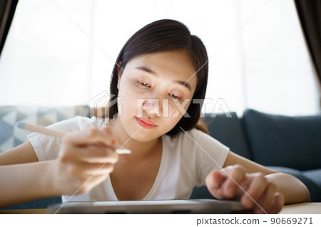 Happy Asian young woman relaxing in the living room and watching a contents and chatting with her friends and drawing an illustration on tablet. Woman enjoy watching video and drawing on tablet. Happy Asian young woman relaxing in the living room and watching a contents and chatting with her friends and drawing an illustration on tablet. Woman enjoy watching video and drawing on tablet. 90669271