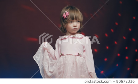 Cute little girl in a vintage pink dress stands on a dark background with a color music. The concept of children's theater productions and developing acting circles Cute little girl in a vintage pink dress stands on a dark background with a color music. The concept of children's theater productions and developing acting circles 90672336