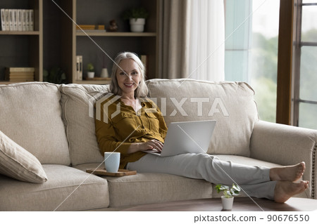 Older woman relaxing on couch with laptop looks at camera Older woman relaxing on couch with laptop looks at camera 90676550