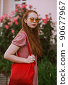 Young beautiful red-haired long-haired woman in a summer pink dress walks through the streets of a summer city. 90677967