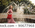 Young beautiful red-haired long-haired woman in a summer pink dress walks through the streets of a summer city. 90677968