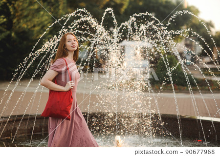 Young beautiful red-haired long-haired woman in a summer pink dress walks through the streets of a summer city. Young beautiful red-haired long-haired woman in a summer pink dress walks through the streets of a summer city. 90677968
