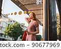 Young beautiful red-haired long-haired woman in a summer pink dress walks through the streets of a summer city. 90677969