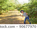A cheerful kid in jeans, a T-shirt and a blue hat is playing in the garden among the apple trees. 90677970