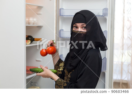 A Muslim woman in national black clothes takes vegetables from the refrigerator for salad 90683466