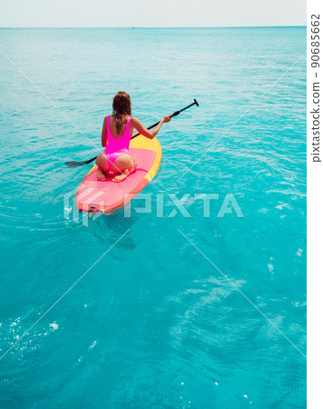 Attractive woman in pink swimwear floating on stand up paddle board on a quiet ocean. SUP surfing with woman in tropical sea Attractive woman in pink swimwear floating on stand up paddle board on a quiet ocean. SUP surfing with woman in tropical sea 90685662