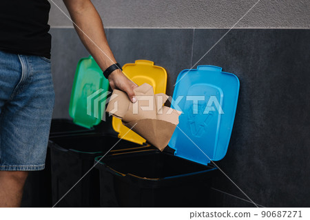 Close-up of man throwing peace of cardboard in recycling bins. Humen hand holding paper near the plastic bin Close-up of man throwing peace of cardboard in recycling bins. Humen hand holding paper near the plastic bin 90687271