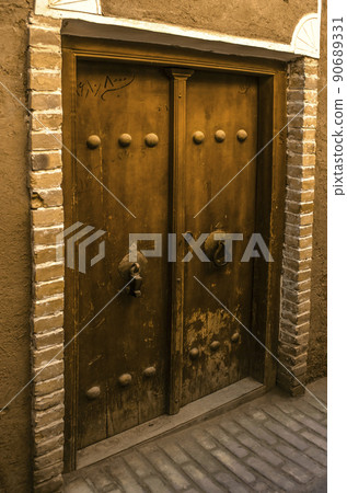 An old double-leaf wooden front door framed with brick on the wall of an adobe house in the ancient adobe city of Yazd. 90689331