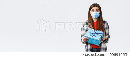 Covid-19, lifestyle, holidays and celebration concept. Excited cute birthday girl holding wrapped box, curious what inside, receive gift, wearing medical mask to prevent coronavirus outbreak 90691965