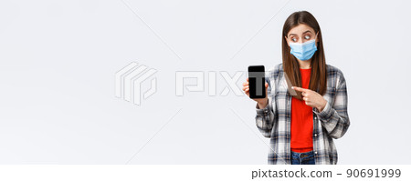 Different emotions, covid-19, social distancing and technology concept. Excited and enthusiastic good-looking girl in medical mask, pointing looking at mobile phone display, promote app 90691999
