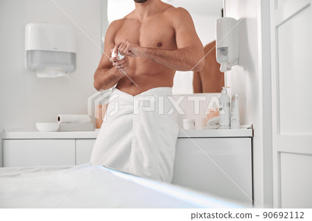 Athletic man with bare chest and towel on hips holds cream leaning onto cabinet in bathroom Athletic man with bare chest and towel on hips holds cream leaning onto cabinet in bathroom 90692112