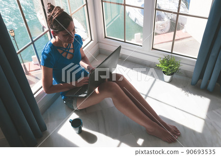 Young woman working with laptop at the window at home over the marina. Freelance lifestyle and work at home concept. 90693535