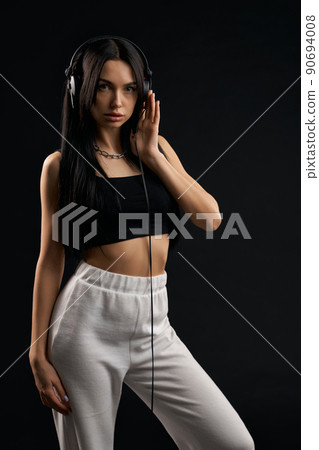 Woman wearing fashion sport clothes listening music with headphones inside. Front view of pretty female model in black top posing with earphones, isolated on black background. Concept of fashion. 90694008