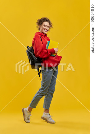 Female student with backpack and books, standing on tiptoe, shyly bent one leg in studio. Front view of cute curly girl looking at camera, smiling, isolated on orange background. Concept of studying. Female student with backpack and books, standing on tiptoe, shyly bent one leg in studio. Front view of cute curly girl looking at camera, smiling, isolated on orange background. Concept of studying. 90694010