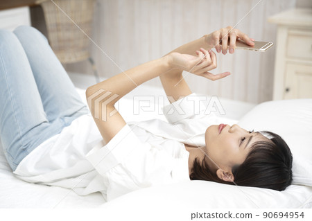 一名年輕女子仰面躺在床上,操作著她的智能手機 一名年輕女子仰面躺在床上,操作著她的智能手機 90694954