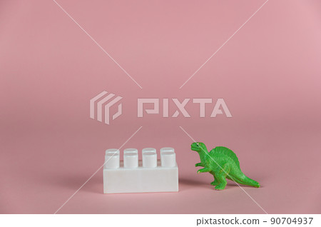 A dinosaur miniature and a white block of children's constructor against a pink background. Small green miniature of a predatory dinosaur. Close-up. Selective focus. A dinosaur miniature and a white block of children's constructor against a pink background. Small green miniature of a predatory dinosaur. Close-up. Selective focus. 90704937