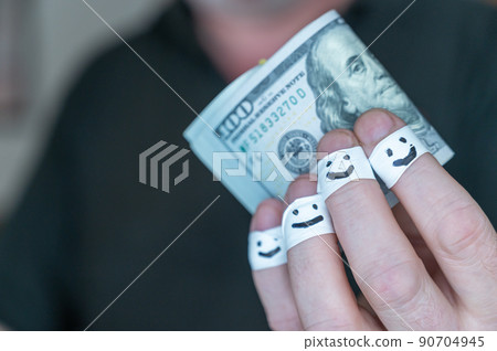 An adult man holds out his hand with $100 bills to the camera. The first phalanges of his fingers are wrapped in white ribbon. Smiling faces are painted on the bandages. Positive emotion concept. An adult man holds out his hand with $100 bills to the camera. The first phalanges of his fingers are wrapped in white ribbon. Smiling faces are painted on the bandages. Positive emotion concept. 90704945