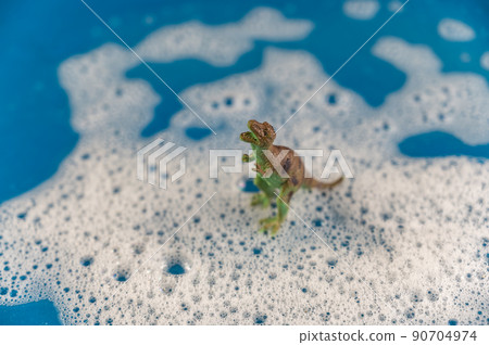 The dinosaur figurine stands in soapy foam. Green predator against a colored background. Bubbles of white detergent foam on the blue surface. Shampoo foam texture. Selective focus. The dinosaur figurine stands in soapy foam. Green predator against a colored background. Bubbles of white detergent foam on the blue surface. Shampoo foam texture. Selective focus. 90704974