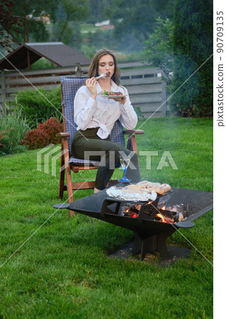Young woman bites grilled meat sitting near barbecue 90709135