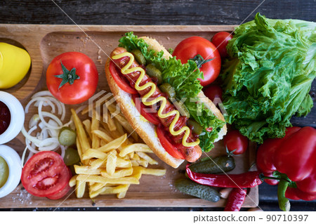 BBQ hot dog served with vegetables and French fries and sauces on wooden serving board BBQ hot dog served with vegetables and French fries and sauces on wooden serving board 90710397