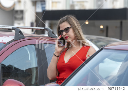 Young beautiful blond woman in red dress has parked her car and is talking on the phone. Modern business woman. Young beautiful blond woman in red dress has parked her car and is talking on the phone. Modern business woman. 90711642
