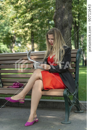 Beautiful young woman in red dress, reading newspaper on bench in park. Attractive girl in park. 90711643