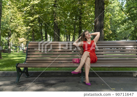 Young woman in red dress sits on bench in summer park. Girl rests in green park. Sunny day. Young woman in red dress sits on bench in summer park. Girl rests in green park. Sunny day. 90711647