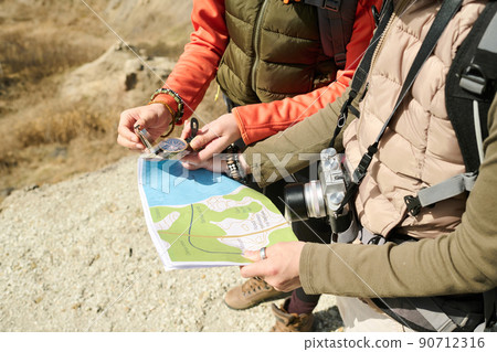 Unrecognizable female tourists wearing backpacks hiking together on sunny spring day reading paper map and using compass to find direction Unrecognizable female tourists wearing backpacks hiking together on sunny spring day reading paper map and using compass to find direction 90712316