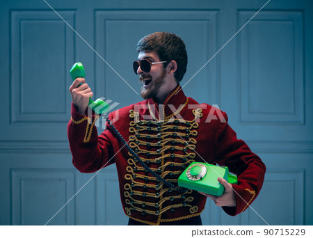 Stylish young man in image of medieval hussar using retro phone isolated on dark blue background. Vintage style, comparison of eras concept. 90715229