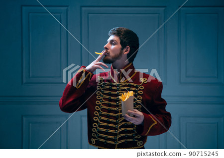 Comic portrait of young man in image of medieval hussar smoking fries isolated on dark blue background. Retro style, comparison of eras concept. 90715245