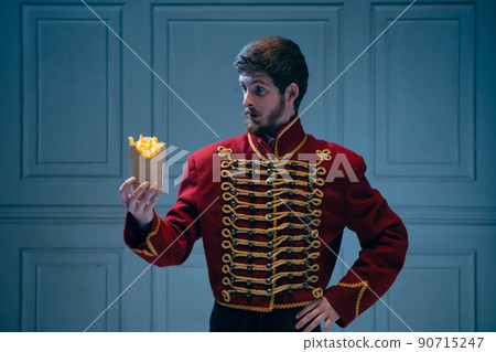 Portrait of young surprised man in image of medieval hussar looking at fries isolated on dark blue background. Retro style, comparison of eras concept. 90715247