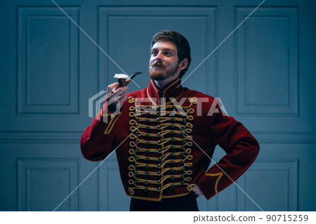 Creative portrait of young man in image of medieval hussar posing isolated on dark blue background. Retro style, comparison of eras concept. Creative portrait of young man in image of medieval hussar posing isolated on dark blue background. Retro style, comparison of eras concept. 90715259