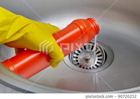 Person's hand in a yellow rubber glove pours pipe cleaner down the drain of a metal kitchen sink 90720252