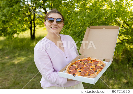 Happy young woman holding hot pizza in box on nature background. Attractive girl feels hungry and loves the taste. Entire pizza for myself. Fast food banner 90721630