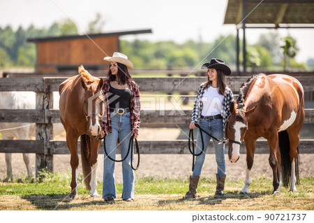 Young women with brown horses stand on a farm looking to the side, smiling Young women with brown horses stand on a farm looking to the side, smiling 90721737