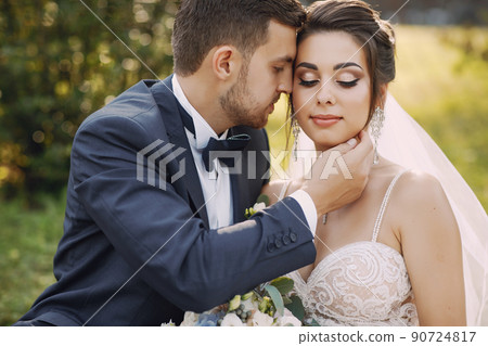 A young and beautiful bride and her husband is standing in a summer park with bouquet of flowers A young and beautiful bride and her husband is standing in a summer park with bouquet of flowers 90724817