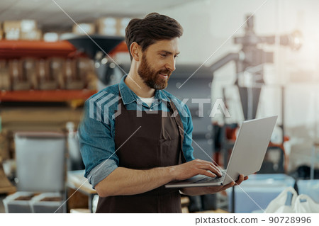 Business owner of small coffee roasting factory working on his workplace using laptop Business owner of small coffee roasting factory working on his workplace using laptop 90728996
