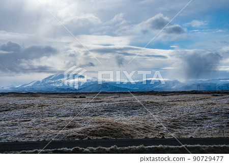 View of road by grassy landscape and snow covered mountains against cloudy sky 90729477
