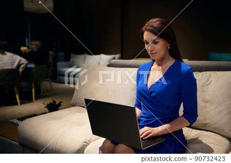 Confident European woman wearing casual dress, sitting on a sofa, planning new business and design project, working on housing project on laptop in a furniture design studio 90732423