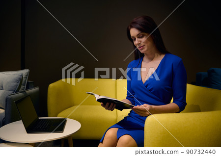 Pretty woman, elegantly dressed in casual blue dress, sitting on yellow couch and leafing through a catalog, choosing new sofa in a furniture design showroom 90732440