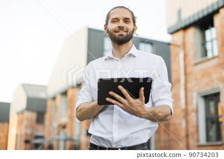 Handsome smiling business man with beard, in white shirt walks around city and using tablet. Handsome smiling business man with beard, in white shirt walks around city and using tablet. 90734203