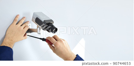 Close up of hands of male worker who uses screwdriver to mount cctv camera on white wall. 90744653