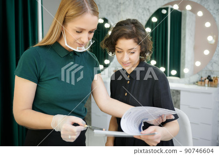 Female makeup artist in protective mask holding pencil and smiling while showing papers to client. Young woman and beauty specialist studying documents in visage studio. 90746604