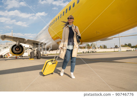 Passenger woman in sunglasses with luggage is outdoors near plane at airport looking away Passenger woman in sunglasses with luggage is outdoors near plane at airport looking away 90746776