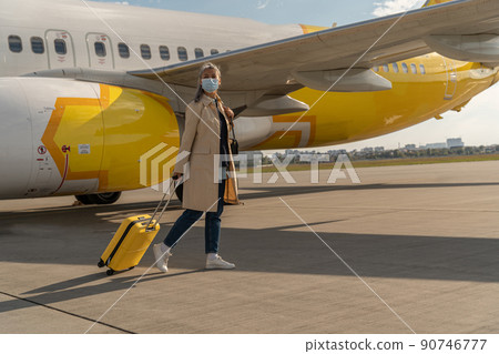Passenger woman in mask with luggage is outdoors near plane looking away Passenger woman in mask with luggage is outdoors near plane looking away 90746777