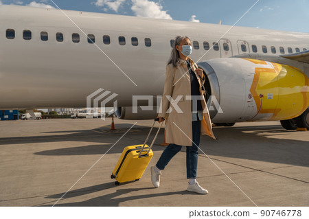 Passenger woman in mask with luggage is outdoors near plane looking away Passenger woman in mask with luggage is outdoors near plane looking away 90746778