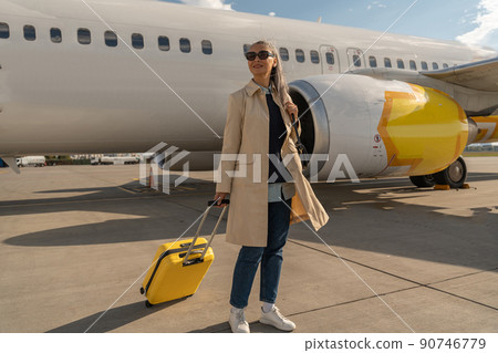Passenger woman in sunglasses with luggage is outdoors near plane at airport looking away Passenger woman in sunglasses with luggage is outdoors near plane at airport looking away 90746779