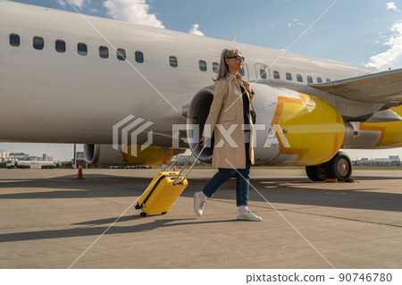 Passenger woman in sunglasses with luggage is outdoors near plane at airport looking away Passenger woman in sunglasses with luggage is outdoors near plane at airport looking away 90746780