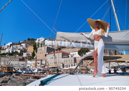 Beautiful young blond woman standing on catamaran at sunny summer day at Hydra marina bay, Greece Beautiful young blond woman standing on catamaran at sunny summer day at Hydra marina bay, Greece 90749204